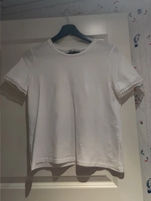 Vit t-shirt med spets från Zara - Superfin vit t-shirt från Zara i mjuk bomull. Klassisk rund hals och korta ärmar med snygg spetskant som ger en extra touch. Perfekt till jeans eller kjol för en clean look. Enkel och stilren design som passar till allt.