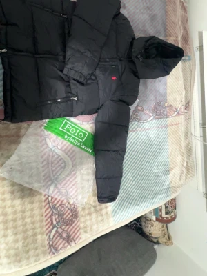 Svart pufferjacka från Polo Ralph Lauren - Svart pufferjacka från Polo Ralph Lauren med huva och röda broderade loggan på bröstet. Jackan har dragkedjor på fickorna och är tillverkad i syntetmaterial. Perfekt för kalla dagar och har en clean, stilren look.