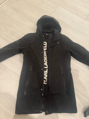 Svart parkajacka från Karl Lagerfeld - Svart parkajacka från Karl Lagerfeld Kids med huva och långa ärmar. Jackan har dragkedja och tryckknappar framtill, samt två vertikala fickor med dragkedja. Insidan har ett mönstrat foder och logotypen syns tydligt på insidan. Perfekt för kyliga dagar.