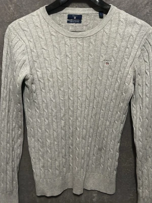 Grå kabelstickad tröja från Gant - Säljer en grå kabelstickad tröja från Gant med rund halsringning och diskret logga på bröstet. Tröjan är tillverkad i stretchig bomull och har långa ärmar samt ribbade muddar vid ärmslut och nederkant. Perfekt för dig som gillar klassisk och stilren look.