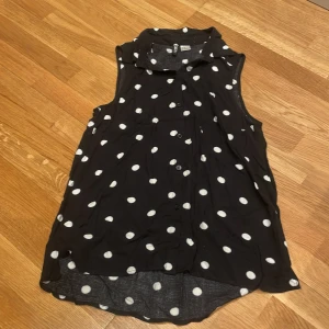 Svart prickig blus från H&M Divided - Säljer en svart ärmlös blus från H&M Divided i storlek 32. Blusen har vita prickar, klassisk krage och knäppning framtill. Lätt och luftig modell som passar perfekt till sommaren. Materialet är väldigt mjukt och skönt