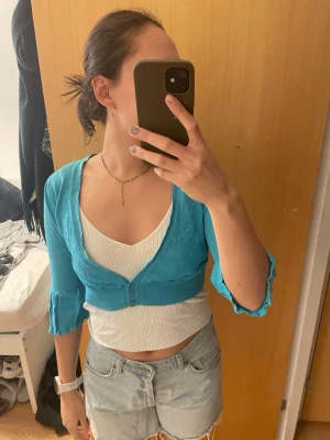 Turkos croppad kofta med volangärm - Säljer en turkos croppad kofta med trekvartsärm och volangdetaljer vid ärmsluten. Koftan har knappar framtill och är i ett mjukt, stickat material. Perfekt att slänga över ett linne eller klänning för en färgklick i outfiten. Vintage från ginatricot passar storlek S/M