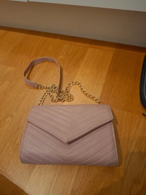 Beige/rosa vintage axelväska med kedja - Snygg beige/rosa axelväska i syntetmaterial med quiltad design och guldfärgad kedja som axelrem. Väskan har ett praktiskt fack med dragkedja inuti och stängs med magnetlås. Perfekt storlek för mobil, plånbok och småsaker. Vintage design. 