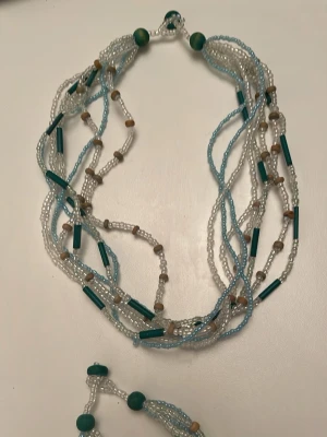 Pärlset halsband och armband blå/vit - Säljer ett pärlset med halsband och armband i flera rader. Pärlorna är i blått, vitt och beige med inslag av avlånga gröna pärlor och små träpärlor. Snyggt boho-inspirerat och perfekt för att lyfta din outfit.