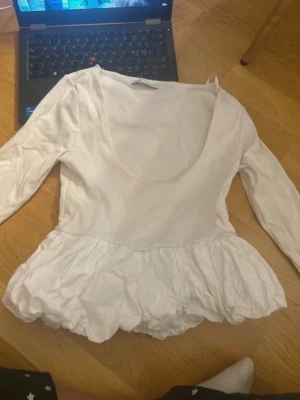 Vit volangblus från Zara - Använd 2 gånger och behöver tvättas innan ny användning! Supersöt vit blus från Zara i storlek M. Den har en djup rundad urringning, långa ärmar och en bred volang nedtill som ger en riktigt fin siluett. Perfekt att styla med jeans eller kjol för en trendig look.