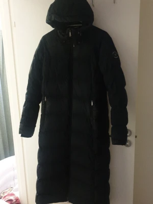 Áhkka Dun kappa. Stl 40 mer som 36 - Lång svart dunjacka med huva och dragkedja framtill. Knappat på sidorna nervvid benen .xJackan har quiltad design, två sidofickor med dragkedja och en diskret logga på ärmen. Perfekt för kalla dagar och riktigt skön att bära. Passar dig som vill ha en varm och stilren vinterjacka. Nypris 3000 