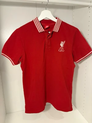 Röd Liverpool FC pikétröja S - Säljer en röd pikétröja från Liverpool FC med vit broderad logga på bröstet. Tröjan har vit-randig krage och ärmslut samt knappar vid halsen. Tillverkad i 100% bomull och har klassisk passform. Perfekt för dig som älskar fotboll och vill visa ditt lag.