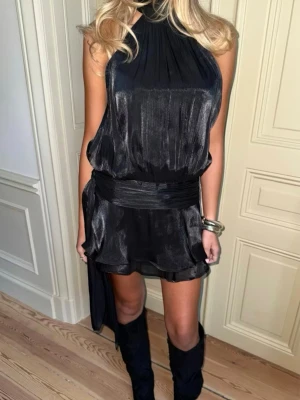 Svart omlottklänning i satin - Supersnygg svart kortklänning i glansigt satinliknande material med hög hals och omlottdetalj i midjan. Klänningen har volangkant nertill, knytning i nacken och är ärmlös. Perfekt för dig som vill sticka ut med en elegant och trendig look.