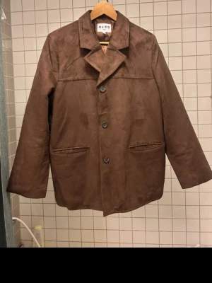 Brun HELT NY jacka i storlek 40 från NA-KD - Säljer en helt ny brun kavaj/jacka i mockaimitation från NA-KD. Jackan har klassisk krage, tre knappar framtill och två fickor. Materialet är mjukt och består av polyester och elastan. Perfekt för dig som vill ha en stilren och trendig look.