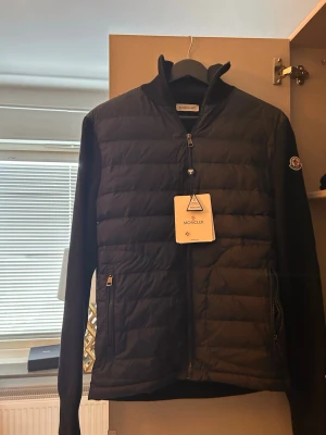 Svart Moncler cardigan herr - Säljer en svart/ mörkblå cardigan från Moncler med quiltad front och släta ärmar. Cardiganen har dragkedja framtill, ståkrage och två fickor med dragkedja. Klassisk Moncler-logga på ärmen och lätt vadderad för extra värme. Perfekt för dig som vill ha en clean och stilren look. Pris kan diskuteras🤗