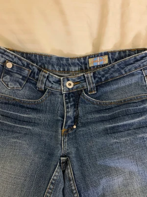 Blå bootcut jeans low waist - Säljer ett par low waist jeans med bootcut passform. Perfekta för dig som gillar retrovibbar och vill ha ett par jeans som sticker ut. Knappen där framme är lös men går enkelt att limma på med lim. 