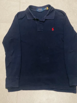 Ralph lauren långärmad piké - Snygg mörkblå långärmad skjorta från Polo Ralph Lauren i klassisk passform. Skjortan har krage, knappar vid halsen och den ikoniska röda Polo-loggan broderad på bröstet. Tillverkad i 100% bomull för en skön känsla.