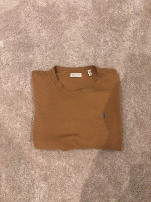Stickad tröja från GANT - Snygg brun stickad tröja från GANT med rund halsringning och diskret broderad logga på bröstet. Perfekt för dig som gillar klassisk och clean stil. Storlek 176cm/16år