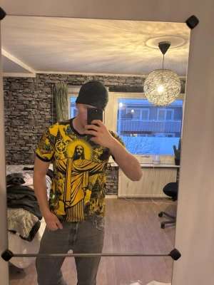 Gul och svart Brasilien t-shirt - Fet t-shirt med stor Kristusstaty och Brasilien-tema i gult och svart. Trycket är detaljerat med motiv av byggnader, änglar och brasilianska flaggor. Svart krage och ärmslut. Perfekt för dig som vill sticka ut med en unik look.