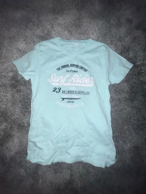 Ljusblå Surf Rider t-shirt från Name It - Snygg ljusblå t-shirt från Name It med tryck 'Surf Rider' och surf-tema framtill. Tillverkad i mjuk ekologisk bomull och har klassisk rund hals och korta ärmar. Perfekt för dig som gillar chill surfstil och vill ha något bekvämt och stilrent.