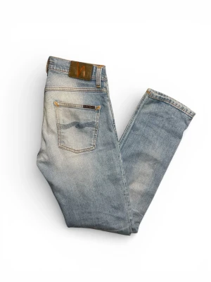Ljusblå Nudie Jeans grim trim 28/32 - Säljer ett par ljusblå slim fit jeans från Nudie Jeans med klassisk femficksdesign och orangea sömmar. Jeansen har en snygg tvättad look och raka ben. Tillverkade i bomull för en skön känsla och stilren passform. Modellen är Grim trim. Jeansen är ljusblåa med en vit tvätt