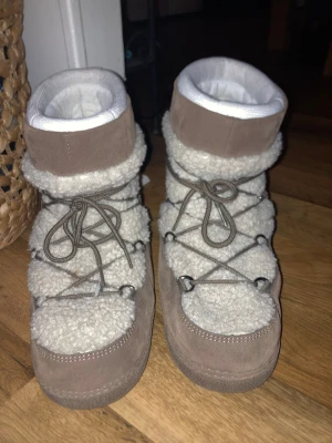 Beige moon boots med snörning - Säljer ett par beige moon boots med fluffig vit teddy och snygga mockadetaljer. Skorna har grov snörning runt hela skon och en mjuk, vadderad insida. Perfekta för kalla vinterdagar och ger en riktigt cozy vibe.