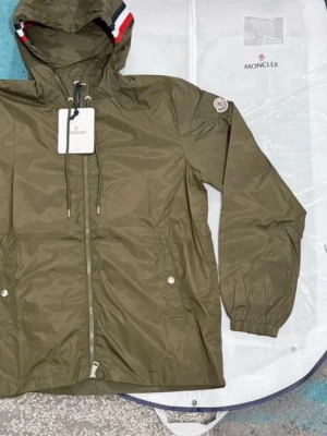 Olivgrön vindjacka från Moncler - Säljer en olivgrön vindjacka från Moncler med huva och snygga detaljer i rött, vitt och blått på huvkanten. Jackan har dragkedja framtill, två fickor med tryckknappar och Moncler-logga på ärmen. Perfekt för dig som vill ha en clean och sportig look. Aldrig använd, pris kan diskuteras vid snabb affär 