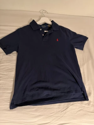 Marinblå pikétröja från Ralph Lauren - Klassisk marinblå pikétröja från Ralph Lauren med röd broderad logga på bröstet. Tröjan har krage, knappar framtill och är kortärmad. Perfekt för en stilren och avslappnad look.