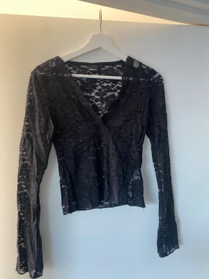 Svart spetsblus från Gina Tricot - Säljer en svart långärmad blus från Gina Tricot i genombruten spets med blommigt mönster. Blusen har omlott-skärning och v-ringad hals, vilket ger en snygg och feminin look. Perfekt att styla med jeans eller kjol för en trendig outfit.
