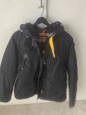 Svart Parajumpers vinterjacka  - Svart vinterjacka från Parajumpers i size M med huva, flera fickor och gul detalj vid kragen. Jackan har patch på ärmen och orange ögla i nacken. Slim fit-modell, perfekt för kalla dagar.