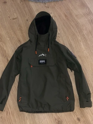 Grön vindjacka från Dope XS - Säljer en grön vindjacka från Dope i storlek XS. Jackan har huva med snörning, stor ficka framtill med Dope-logga och detaljer i orange. Materialet är slitstarkt och perfekt för utomhusaktiviteter. Enkel och clean design med lång ärm och justerbara muddar. strl xs/s
