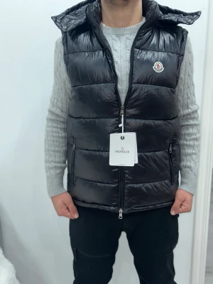 Svart dunväst från Moncler - Säljer en svart, glansig dunväst från Moncler med huva och dragkedja framtill. Västen har Moncler-logga på bröstet och två sidofickor med dragkedja. Perfekt för lager-på-lager och riktigt snygg till höst och vinter.