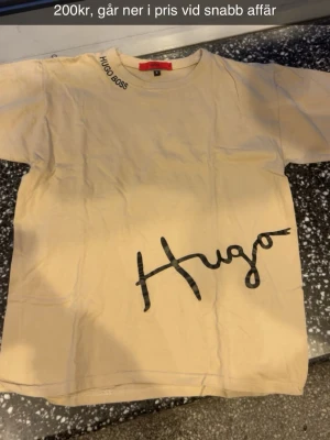 Beige Hugo Boss t-shirt med tryck - Snygg beige t-shirt från Hugo Boss med svart tryck på bröstet där det står 'Hugo' i Cottons signaturstil. På kragen finns även 'Hugo Boss' tryckt i svart. Klassisk rund halsringning och korta ärmar. Perfekt för en clean och stilren look. Går ner i pris vid snabb affär.