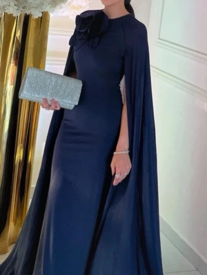 Marinblå långklänning med cape - Säljer en elegant marinblå långklänning med cape och stor dekorativ blomma vid halsen. Klänningen har en slimmad passform och svepande tyg som ger ett dramatiskt intryck. Perfekt för bal eller festliga tillfällen där du vill sticka ut.