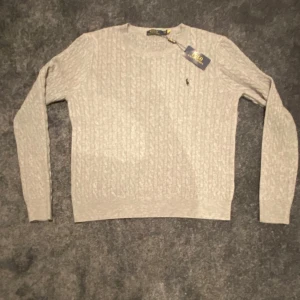 Grå kabelstickad tröja Polo Ralph Lauren - Helt ny kabelstickad grå tröja från Polo Ralph Lauren med rund halsringning och diskret broderad logga på bröstet. Tröjan har lång ärm och ribbade muddar. 
