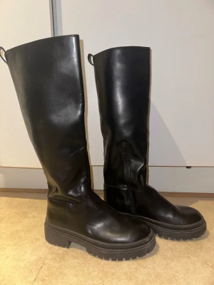 Svarta höga boots i skinn, strl 39 - Säljer ett par svarta höga boots i skinn med grov sula och rund tå. Skorna har dragkedja på insidan och en rejäl platt sula med tydligt mönster undertill. Perfekt för dig som gillar chunky och stilrena boots.