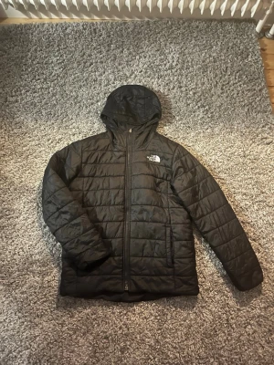 Svart pufferjacka från The North Face - Säljer en svart pufferjacka från The North Face med huva och dragkedja framtill. Jackan har quiltad design och logga på bröstet. Perfekt för kyliga dagar och enkel att matcha med olika outfits.