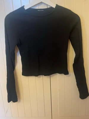 Svart croppad långärmad topp - Säljer en svart croppad topp med långa ärmar och rund hals. Toppen är ribbad och har en tight passform som framhäver midjan. Perfekt att matcha med jeans eller kjol för en stilren look. Väldigt bra skick. Säljer billigt pga växt ut och knappt används.