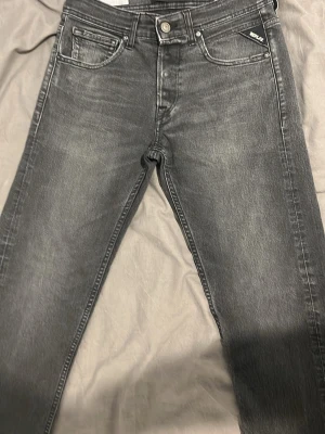 Replay grå med svart jeans med raka ben - Säljer ett par gråa Replay jeans med klassisk femficksdesign och raka ben. Jeansen har snygga slitningar och detaljerade sömmar, samt Replay-logga på fickan och bak vid linningen. Tillverkade i mjukt jeanstyg för skön passform.