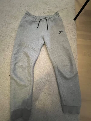 Gråa mjukisbyxor från Nike - Säljer ett par gråa mjukisbyxor från Nike med svart logga på vänster lår. sopass klassiska med resår i midjan och svarta snören för justering. Byxorna har fickor på sidorna och är tillverkade i mjukt bomullsmaterial. Perfekta för chill eller träning.