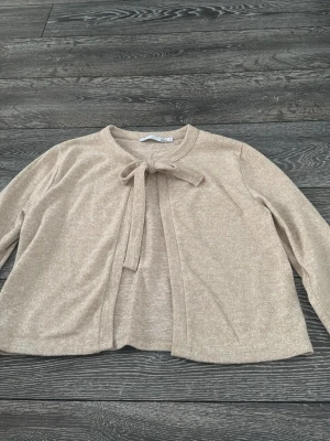 Beige kofta med knyt från 157 - Mysig beige kofta från 157 med knytband vid halsen. Koftan har lång ärm och är tillverkad i ett mjukt material som känns skönt mot huden. Perfekt att slänga på sig över en topp för en chill look.