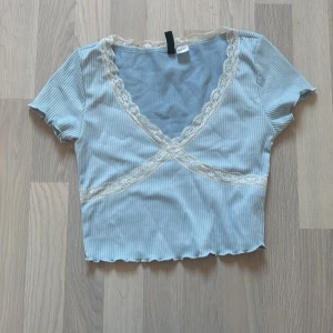 Ljusblå croppad ribbad topp med spets - Superfin ljusblå croppad topp med ribbat tyg och korta ärmar. V-ringad omlott med vit spetskant runt halsen och över bysten. Söt volangkant nertill och figurnära passform. Perfekt för dig som gillar en feminin och trendig stil.