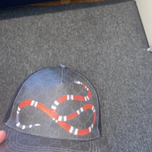 Gucci keps med ormtryck - Snygg svart keps från Gucci med mesh på baksidan och justerbar rem. Framsidan har ett coolt rött och vitt ormtryck ovanpå det klassiska GG-mönstret. Materialet är en mix av polyester och bomull, perfekt för dig som vill sticka ut med designerstil.