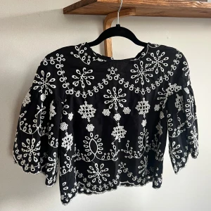 broderad blus - Superfin svart blus med vita broderade blommönster och vida trekvartsärmar. Toppen är croppad med vågig kant nertill och har en rund halsringning. 