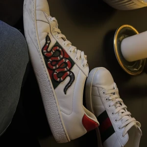 Gucci ace snake - Säljer ett par vita Gucci Ace sneakers i skinn med klassiska gröna och röda ränder på sidan. Ena skon har en broderad orm i rött, svart och vitt. Båda har metallicröd detalj på hälen och vita snören. Snygg och ikonisk modell för dig som vill sticka ut.