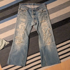 Levis 501 ljusblå slitna jeans - Ljusblå Levis jeans med rejält slitna och trasiga detaljer framtill och på bakfickorna. Klassisk femficksmodell. Jeansen har en cool vintagekänsla och är tillverkade i robust denim.