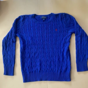 Blå kabelstickad tröja Polo Ralph Lauren - Kabelstickad blå tröja från Polo Ralph Lauren med rund halsringning och diskret röd logga broderad på bröstet. Tröjan har långa ärmar och ribbade muddar vid ärmslut och nederkant. Perfekt för dig som gillar klassisk och stilren look.