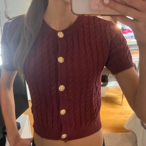 Vinröd stickad kofta från H&M - Vinröd stickad kofta från H&M med korta ärmar och snygga guldknappar framtill. Kabelstickat mönster och ribbade kanter ger en klassisk vibe. Perfekt till lager-på-lager och funkar året runt. Croppad passform som sitter snyggt till både jeans och kjol.