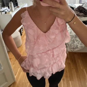 Rosa volangig linnetopp med blommönster - Supersöt ljusrosa linnetopp med tunna axelband och flera lager volanger. Toppen har ett subtilt blommönster i tyget och v-ringning framtill.  Materialet känns lätt och luftigt.