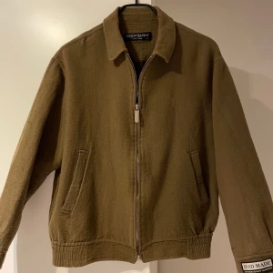 Brun ulljacka från Yves Saint Laurent - Säljer en brun jacka från Yves Saint Laurent i ull med klassisk krage, dragkedja framtill och diskret YSL-brodyr på bröstet. Jackan har två snedställda fickor och ribbade muddar nedtill. Perfekt för dig som gillar stilren och tidlös design.