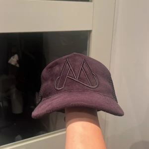 Lila keps med broderad M-logga - Snygg lila keps med stor broderad M-logga framtill. Kepsen har mesh på sidorna för extra ventilation och är tillverkad i ett mjukt material som ger en cool look. Perfekt accessoar för att lyfta din outfit.