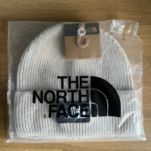 Beige mössa från The North Face - Snygg beige ribbstickad mössa från The North Face med uppvikt kant och klassisk logotyp framtill. Perfekt accessoar för kyliga dagar, tillverkad i mjukt material som håller dig varm och stilren. Leveras i originalförpackning. 