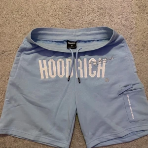 Blå Hoodrich shorts med tryck - Snygga ljusblå shorts från Hoodrich med stort vitt broderat tryck framtill och textdetaljer bak. Modellen har snörning i midjan och en praktisk ficka på sidan. Perfekta för chill dagar och streetwear-vibe. Vinted shorts som inte säljs längre 