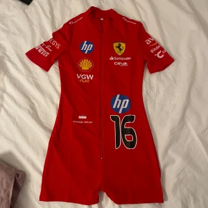Ferrari kort jumpsuit - Sportig röd kort jumpsuit inspirerad av Ferrari och Charles Leclerc, med tryckta loggor från HP, Shell och Santander. Jumpsuiten har dragkedja framtill, korta ärmar och figurnära passform. Aldrig använd!