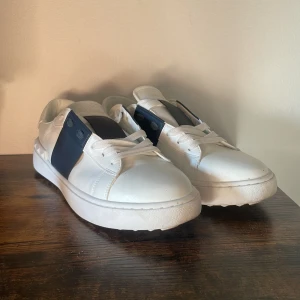 Valentino Garavani vita sneakers - Snygga vita sneakers från Valentino Garavani med bred marinblå detalj på sidan och ikoniska nitar runt hälen. Skorna har snörning, rund tå och är tillverkade i skinn för en lyxig känsla. Perfekta för dig som vill ha en stilren men edgy look.
